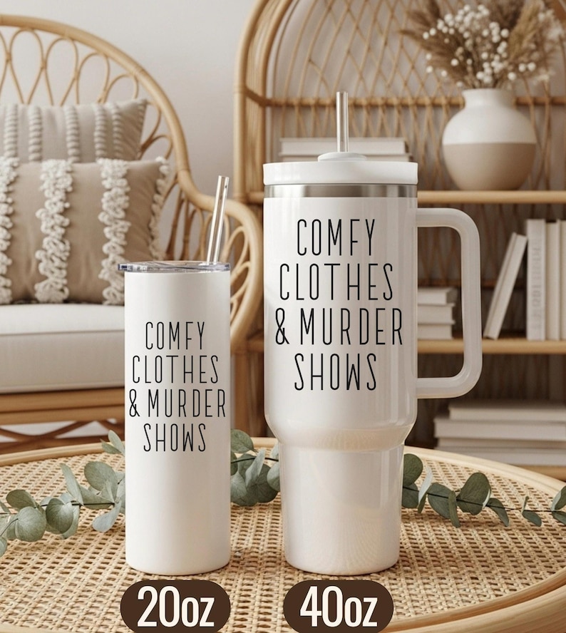 May include: Two white insulated tumblers with the text "COMFY CLOTHES & MURDER SHOWS" in black. One tumbler is 20oz, the other is 40oz. Both have a straw and a lid. The tumblers are on a woven tray.