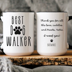 May include: Two white ceramic mugs with black handles and black rims. One mug says "Best Dog Walker" with a paw print. The other mug says "Thank you for all the love, cuddles and treats, Katie. I woof you! Brownie".