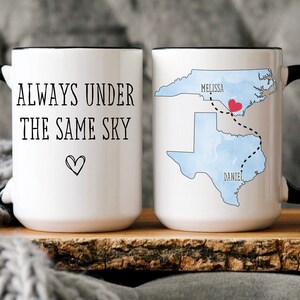 May include: Two white coffee mugs with black handles. One mug has the text "ALWAYS UNDER THE SAME SKY" with a small heart. The other mug has a map of North Carolina and Texas with a red heart in the middle of the states and the text "MELISSA" and "DANIEL" on the map.