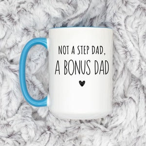 May include: White ceramic mug with a blue handle and interior. The mug features the text "NOT A STEP DAD, A BONUS DAD" in black, with a small black heart below the text. The mug is set against a soft, white and gray textured background.