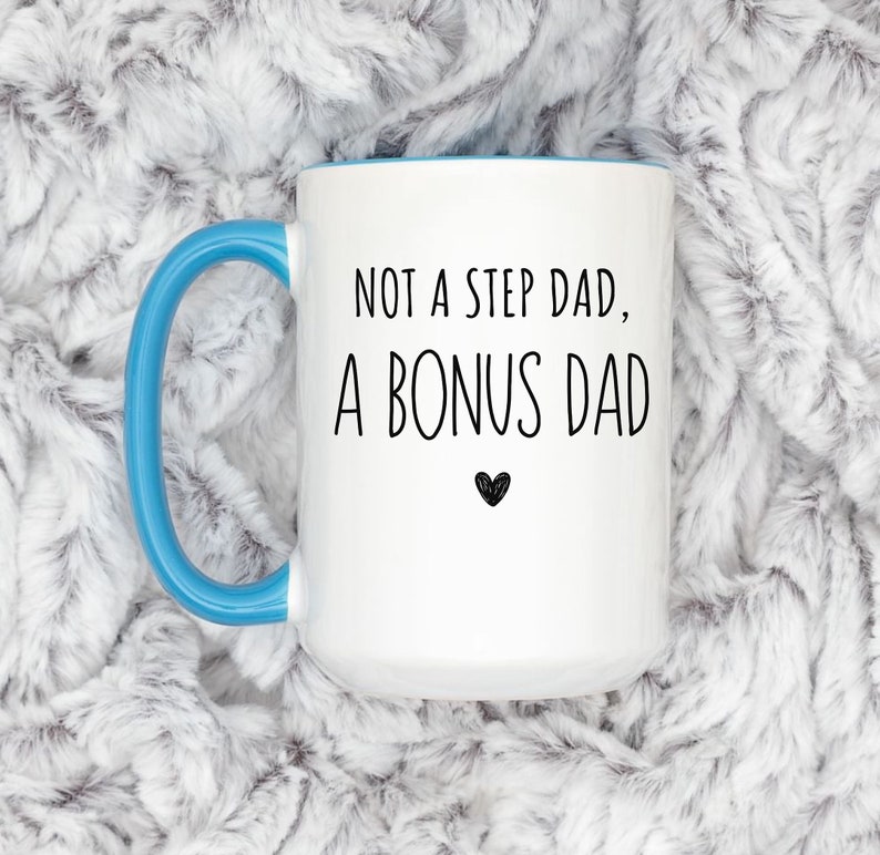 May include: White ceramic mug with a blue handle and interior. The mug features the text "NOT A STEP DAD, A BONUS DAD" in black, with a small black heart below the text. The mug is set against a soft, white and gray textured background.