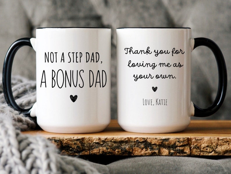 May include: White ceramic mug with black handle and rim. The mug has the text "NOT A STEP DAD, A BONUS DAD" and a heart on one side. The other side says "Thank you for loving me as your own. Love, Katie."