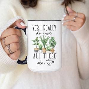 May include: White ceramic mug with a black interior and handle, featuring the text "YES I REALLY do need ALL THESE plants" and a watercolor illustration of various potted plants.