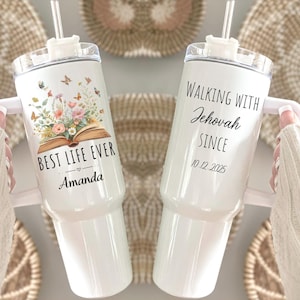 May include: Two white insulated tumblers with handles and straws. One features the text "BEST LIFE EVER" with floral and butterfly graphics, and the name "Amanda." The other says "WALKING WITH Jehovah SINCE 10.12.2025."