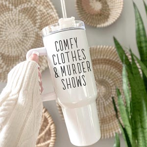 May include: White tumbler with a handle and a clear lid. The tumbler has the text "COMFY CLOTHES & MURDER SHOWS" in black. A white straw is inserted into the lid. The tumbler is held in front of a wall with woven wall decor and a green plant.