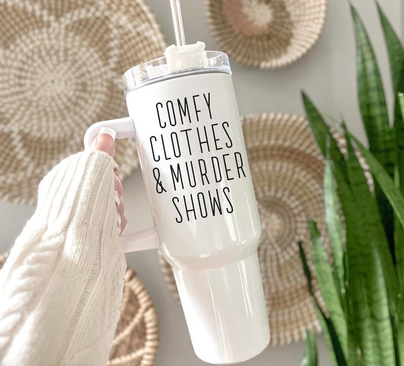 May include: White tumbler with a handle and a clear lid. The tumbler has the text "COMFY CLOTHES & MURDER SHOWS" in black. A white straw is inserted into the lid. The tumbler is held in front of a wall with woven wall decor and a green plant.