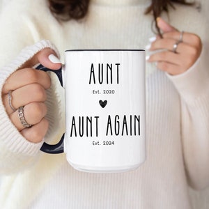 May include: White ceramic mug with a black rim and handle. The mug has black text that reads "Aunt Est. 2020" with a small heart symbol and "Aunt Again Est. 2024".