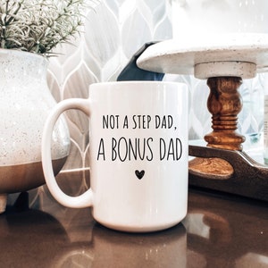 May include: White ceramic mug with the text "NOT A STEP DAD, A BONUS DAD" in black, with a small heart symbol. The mug has a large handle and is sitting on a brown surface.