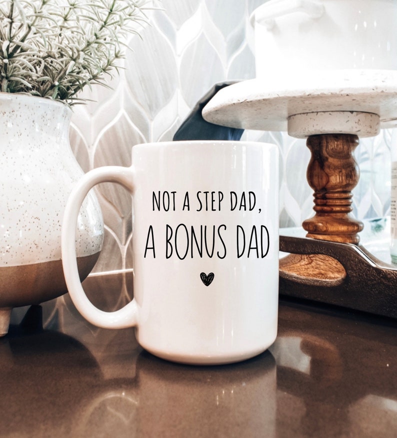 May include: White ceramic mug with the text "NOT A STEP DAD, A BONUS DAD" in black, with a small heart symbol. The mug has a large handle and is sitting on a brown surface.