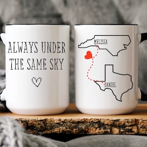 May include: Two white coffee mugs with black handles. One mug says "Always under the same sky" with a small heart. The other mug has a map of the United States with a red heart and a dotted line connecting North Carolina and Texas. The text "Melissa" is above North Carolina and "Daniel" is below Texas.