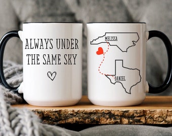Op de afbeelding: Twee witte koffiemokken met zwarte handvatten. Op de ene mok staat "Always under the same sky" met een klein hartje. De andere mok heeft een kaart van de Verenigde Staten met een rood hartje en een stippellijn die North Carolina en Texas verbindt. De tekst "Melissa" staat boven North Carolina en "Daniel" staat onder Texas.