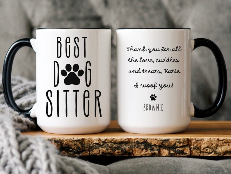 May include: White ceramic coffee mug with a black handle and black rim. The mug has the text "Best Dog Sitter" with a paw print in the middle of the text. The other side of the mug has the text "Thank you for all the love, cuddles and treats, Katie. I woof you! Brownie".
