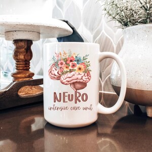 May include: White ceramic mug featuring a detailed illustration of a brain adorned with colorful flowers. The mug has the word "NEURO" and "intensive care unit" printed in a brown font. The mug is on a brown surface.