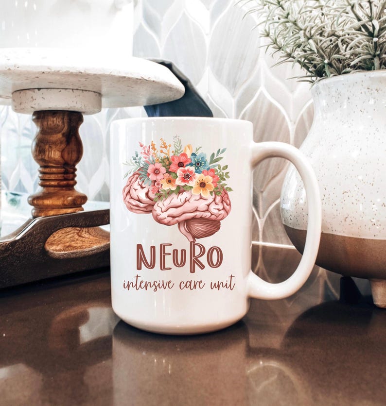 May include: White ceramic mug featuring a detailed illustration of a brain adorned with colorful flowers. The mug has the word "NEURO" and "intensive care unit" printed in a brown font. The mug is on a brown surface.