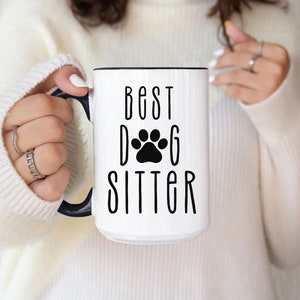 May include: White ceramic mug with a black rim and handle. The mug has black text that reads "Best Dog Sitter" with a black paw print between the words "Dog" and "Sitter".