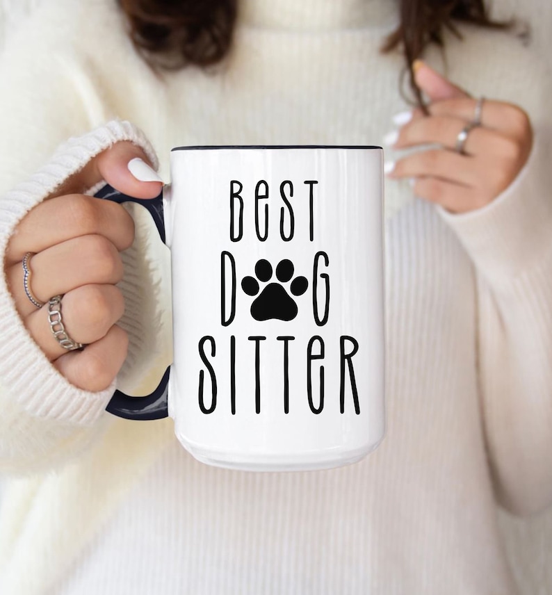 May include: White ceramic mug with a black rim and handle. The mug has black text that reads "Best Dog Sitter" with a black paw print between the words "Dog" and "Sitter".