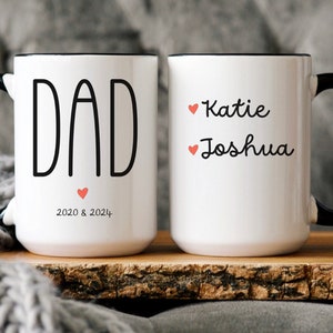 May include: White ceramic mug with a black handle and rim. The mug has the word "DAD" printed in black with a small red heart below. The text "2020 & 2024" is printed below the heart. The other mug is white with a black handle and rim. The text "Katie" and "Joshua" are printed in black with a small red heart before each name.