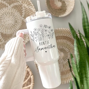 May include: White tumbler with a handle, featuring a floral design and the text "AUNTIE Samantha" in black script. The tumbler includes a clear lid and a straw. The background includes woven wall decor and green plants.