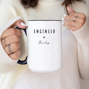 May include: A white ceramic mug with a black interior and handle. The mug features the word "ENGINEER" in black, a small heart, and the name "Emily" in a script font. The mug is held by a person.