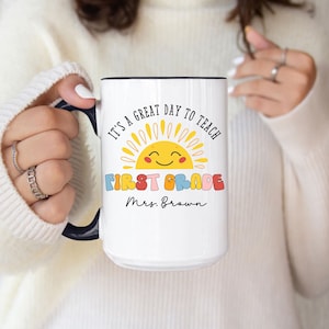 May include: White ceramic mug with a black handle and rim. The mug features the text "IT'S A GREAT DAY TO TEACH FIRST GRADE Mrs. Brown" with a smiling sun graphic in yellow, orange, and blue. A perfect gift for a teacher.