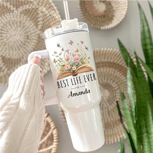 May include: White tumbler with a handle, featuring the text "BEST LIFE EVER" and the name "Amanda". The tumbler is decorated with a floral and butterfly design. It includes a clear lid and a straw.
