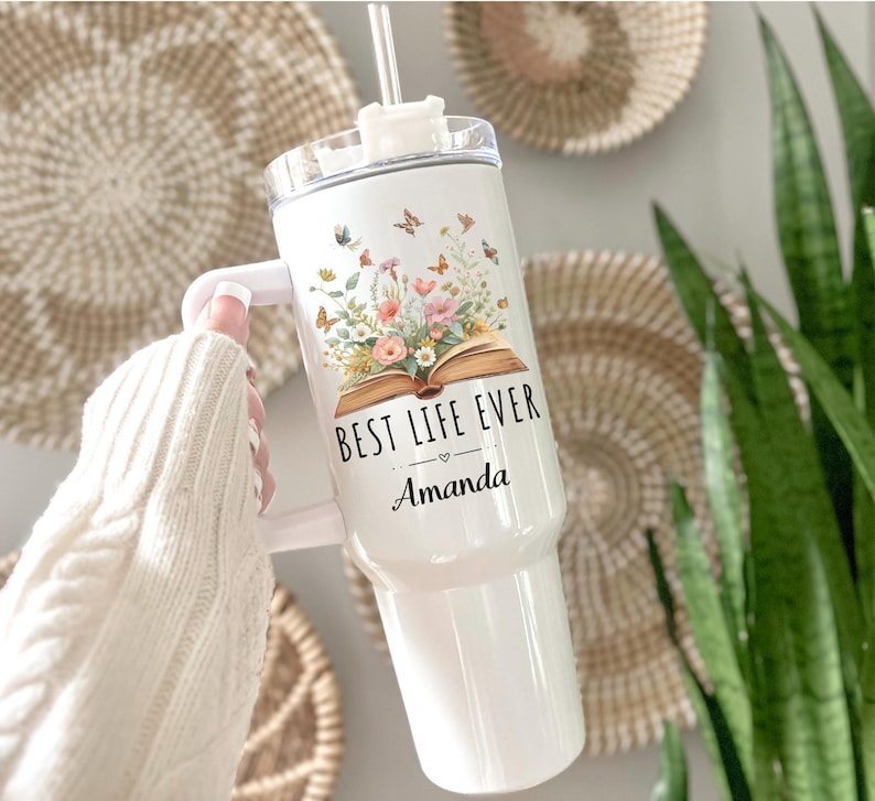May include: White tumbler with a handle, featuring the text "BEST LIFE EVER" and the name "Amanda". The tumbler is decorated with a floral and butterfly design. It includes a clear lid and a straw.