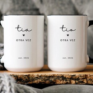 May include: Two white ceramic mugs with black handles and rims. Each mug has the word "tia" above "OTRA VEZ" and "est. 2026" in black script. A small black heart is between the words. The mugs are on a wooden surface.