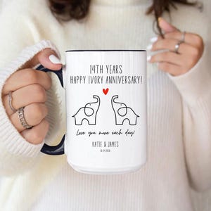 May include: White ceramic mug with a black rim and handle. The mug has a black inscription that reads "14th Years Happy Ivory Anniversary!" with two elephants facing each other and a red heart above them. The inscription continues with "Love you more each day!" and "Katie & James 10.24.2010".