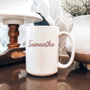 May include: A white ceramic mug with the name "Samantha" in a cursive font. The mug has a large handle and is sitting on a dark brown surface. A decorative vase and a wooden tray are in the background.