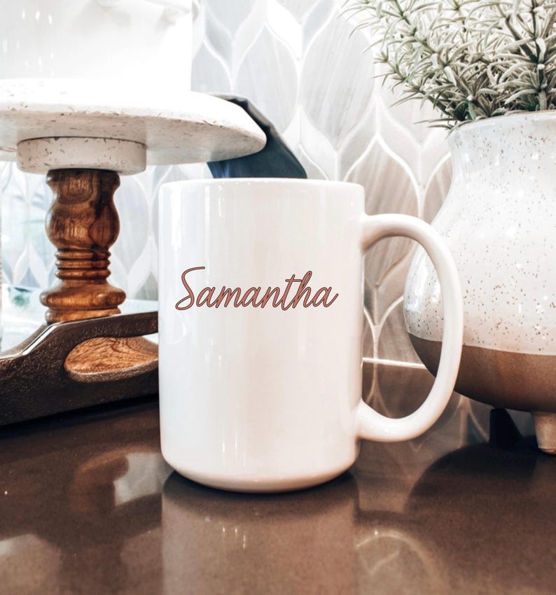 May include: A white ceramic mug with the name "Samantha" in a cursive font. The mug has a large handle and is sitting on a dark brown surface. A decorative vase and a wooden tray are in the background.