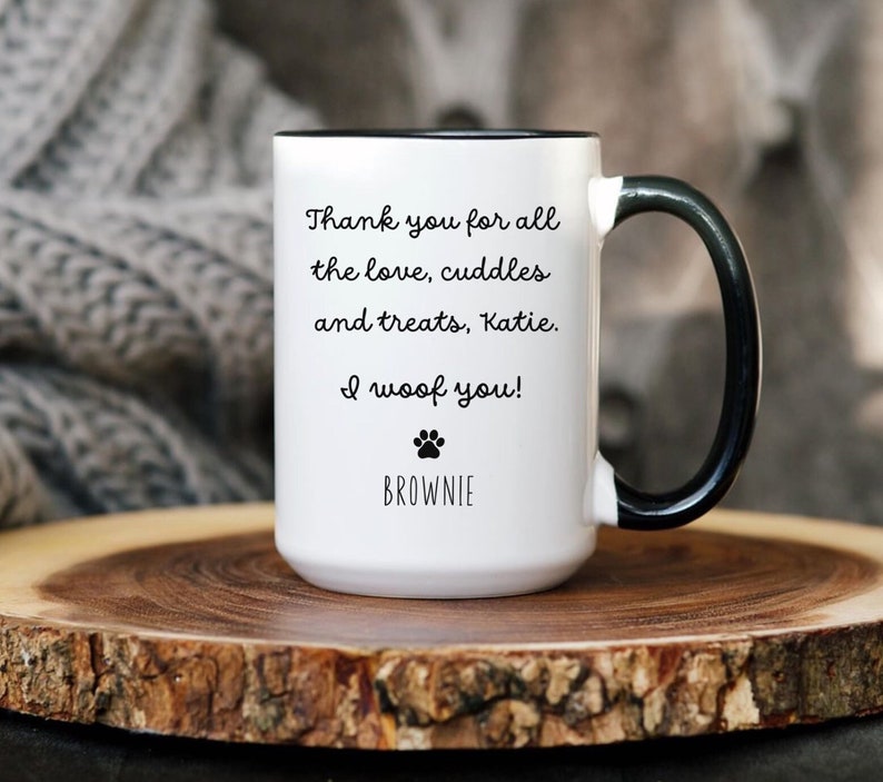 May include: White ceramic mug with a black rim and handle. The mug has a paw print and the text "Thank you for all the love, cuddles and treats, Katie. I woof you! BROWNIE" printed on it.