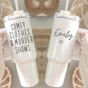 May include: Two white insulated tumblers with handles and straws. One tumbler has the text "COMFY CLOTHES & MURDER SHOWS" in black. The other has the name "Emily" and a heart symbol in black. The tumblers are held against a neutral background.