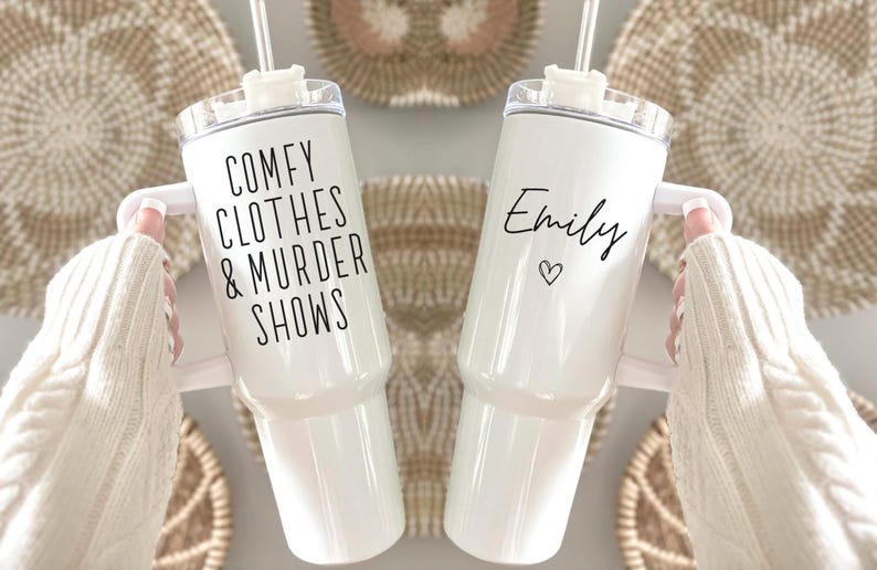 May include: Two white insulated tumblers with handles and straws. One tumbler has the text "COMFY CLOTHES & MURDER SHOWS" in black. The other has the name "Emily" and a heart symbol in black. The tumblers are held against a neutral background.