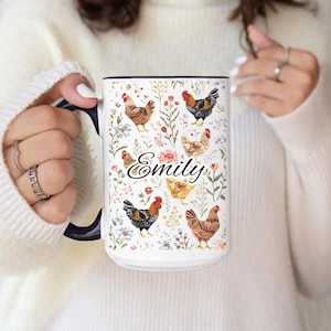 May include: White ceramic mug with a black interior and handle. The mug features a floral design with various chickens and the name "Emily" in cursive. The design includes red, yellow, and brown chickens.
