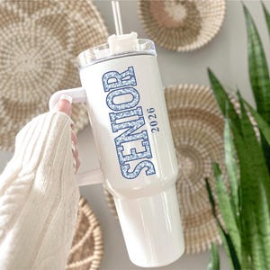 May include: White tumbler with a handle, featuring the word "SENIOR" in blue floral print and the year "2026". The tumbler has a clear lid and a straw. The background includes woven wall decor and greenery.