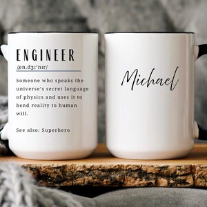 May include: Two white ceramic mugs with black handles and rims. One mug features the word "ENGINEER" with a definition and the phrase "See also: Superhero." The other mug is personalized with the name "Michael" in script.