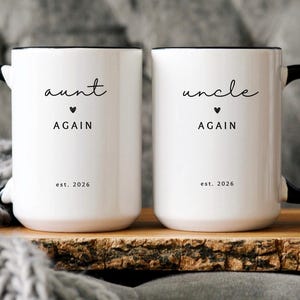 May include: Two white ceramic mugs with black handles and rims. One mug reads "aunt again" with a heart and "est. 2026". The other mug reads "uncle again" with a heart and "est. 2026".