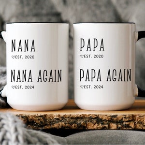 May include: Two white ceramic mugs with black handles and black rims. The mug on the left says "NANA ♡EST. 2020 NANA AGAIN ♡EST. 2024". The mug on the right says "PAPA ♡EST. 2020 PAPA AGAIN ♡EST. 2024".