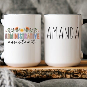 May include: Two white ceramic mugs with black handles. The mug on the left has a floral design and the text "ADMINISTRATIVE assistant" in rainbow colors. The mug on the right has the name "AMANDA" in black text.
