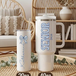 May include: Two white insulated tumblers with lids and straws. One is a 20oz tumbler with a blue bow and the name "Katherine". The other is a 40oz tumbler with "SENIOR 2026" in blue. Both are on a woven surface.
