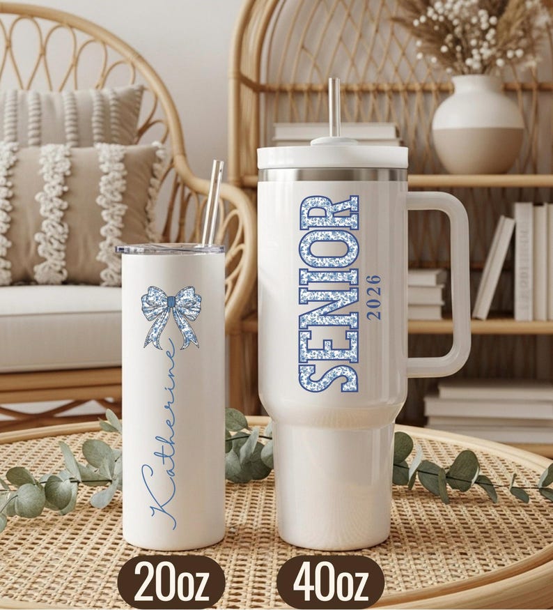 May include: Two white insulated tumblers with lids and straws. One is a 20oz tumbler with a blue bow and the name "Katherine". The other is a 40oz tumbler with "SENIOR 2026" in blue. Both are on a woven surface.