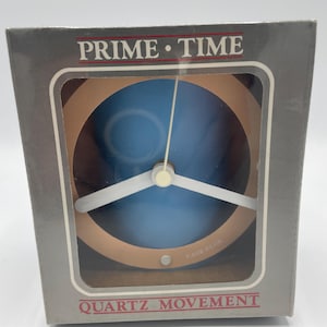 May include: A silver box with a blue and white clock inside. The clock face is surrounded by a brown ring. The box has the words "Prime Time" and "Quartz Movement" printed on it.