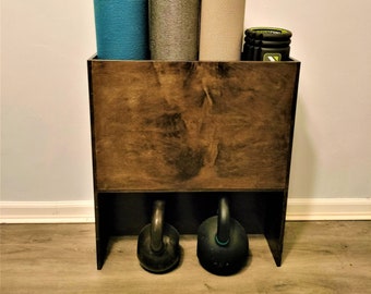 May include: A dark brown wooden storage rack with three compartments for storing yoga mats and other fitness equipment. The rack has a shelf on top and a lower shelf for storing two kettlebells.