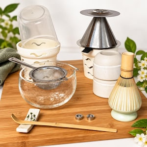 May include: A matcha tea set including a clear glass bowl with a metal mesh strainer, a bamboo whisk on a ceramic stand, a bamboo scoop on a ceramic rest, a metal cone filter, a white ceramic base, and two small metal rings.