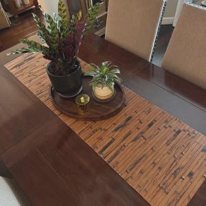 May include: A dark brown wooden dining table with a bamboo table runner. Two beige upholstered chairs are visible. A round wooden tray holds two potted plants and a small gold candle holder.
