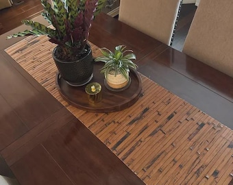 May include: A dark brown wooden dining table with a bamboo table runner. Two beige upholstered chairs are visible. A round wooden tray holds two potted plants and a small gold candle holder.