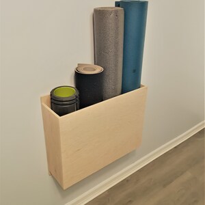 May include: A light wood wall mounted shelf with three rolled up yoga mats. The mats are gray, blue, and black. The shelf is mounted on a white wall.