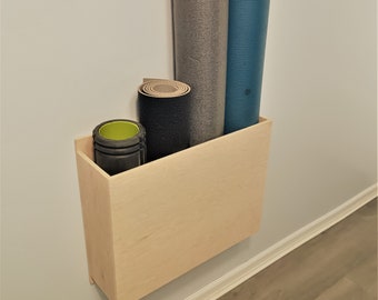 May include: A light wood wall mounted shelf with three rolled up yoga mats. The mats are gray, blue, and black. The shelf is mounted on a white wall.