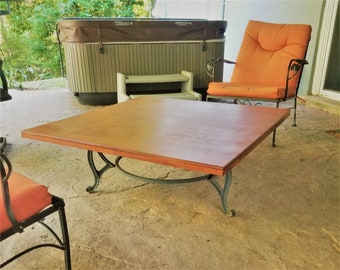 May include: A square wooden coffee table with a brown top and a black metal base. The table is sitting on a patio with a hot tub and a chair in the background.