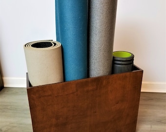 May include: A wooden storage bin filled with rolled-up yoga mats and a foam roller. The mats are in various colors, including teal, gray, and beige. The bin is made of dark brown wood and sits on a light gray floor.